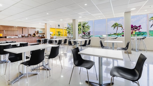 A modern cafeteria with white tables and black chairs, featuring a large window with a view of palm trees and flowers. - City Express by Marriott Tuxtla Gutierrez (Tuxtla Gutierrez)