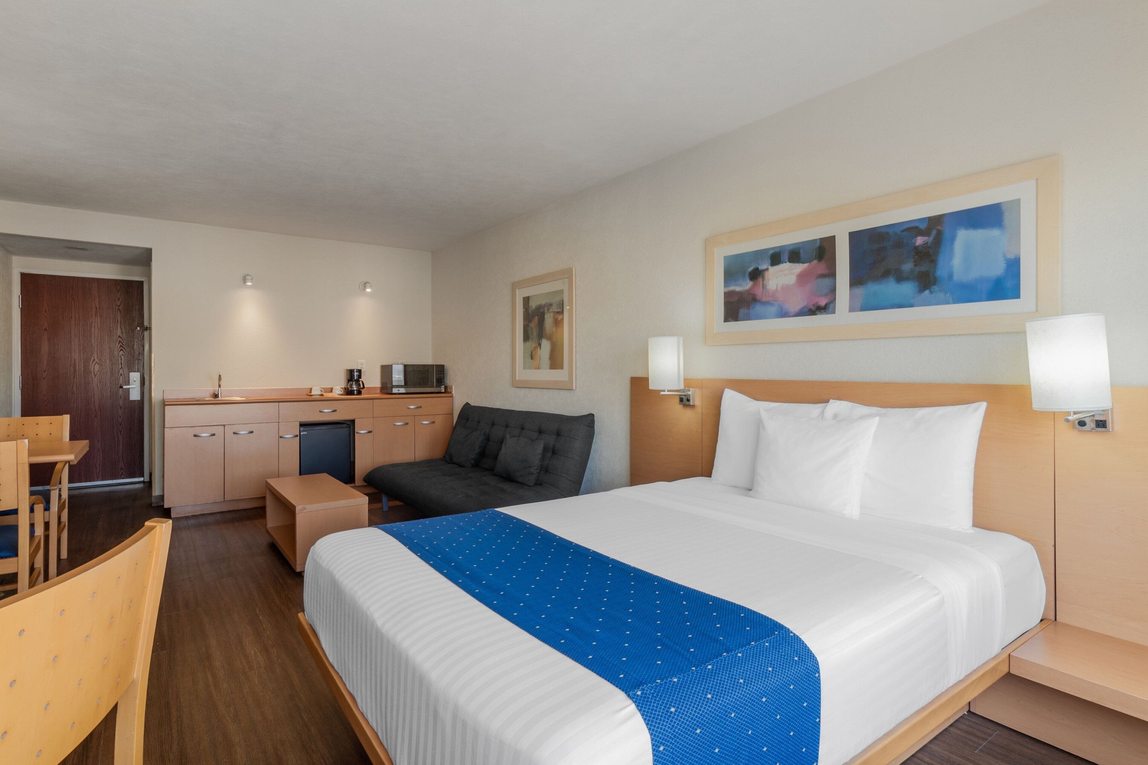 Habitación de hotel con cama, escritorio, sillón, sofá, kitchenette y un cuadro en la pared.