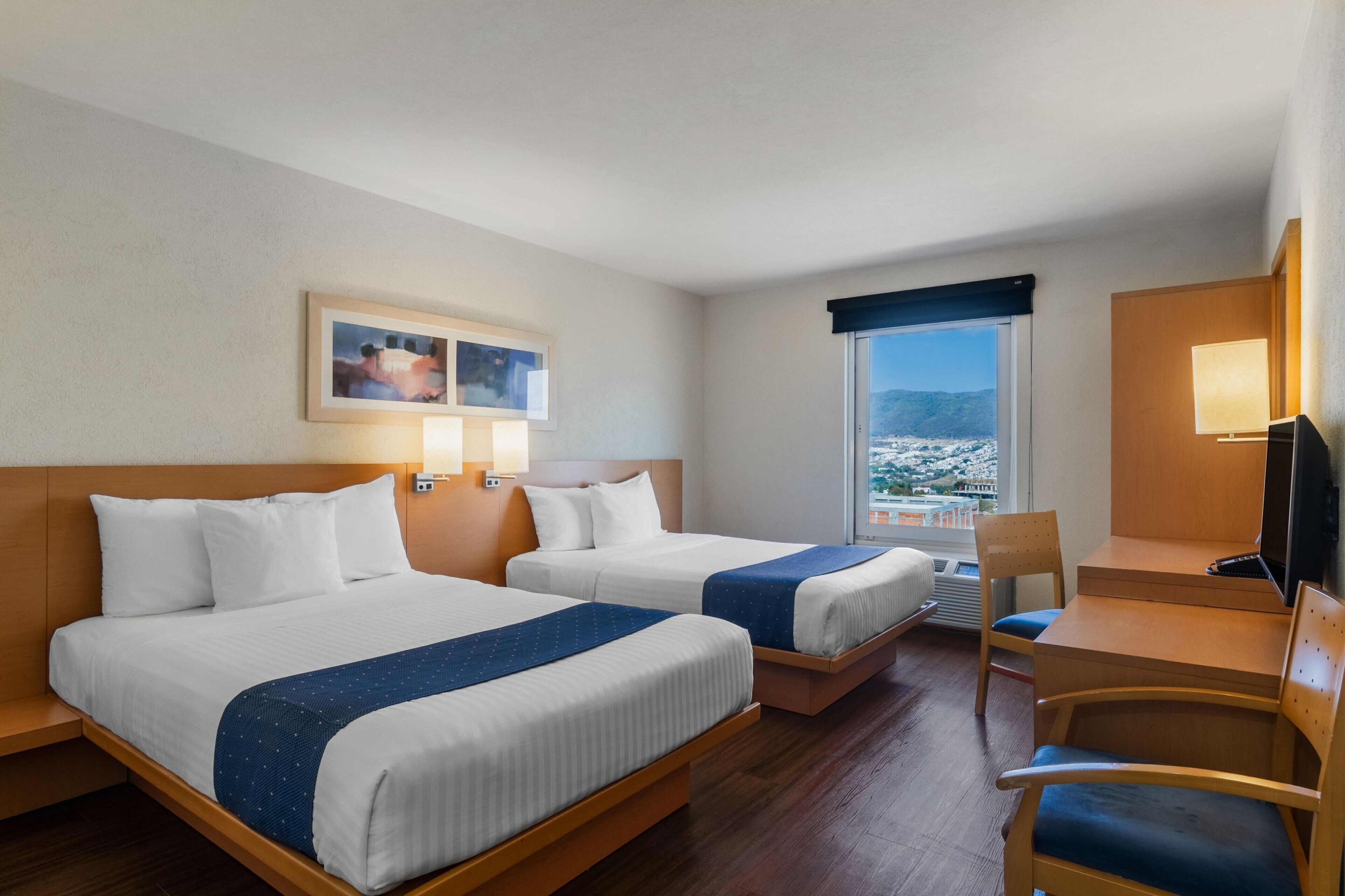 Habitación de hotel con dos camas, un escritorio, una silla, un televisor y ventana con vistas.