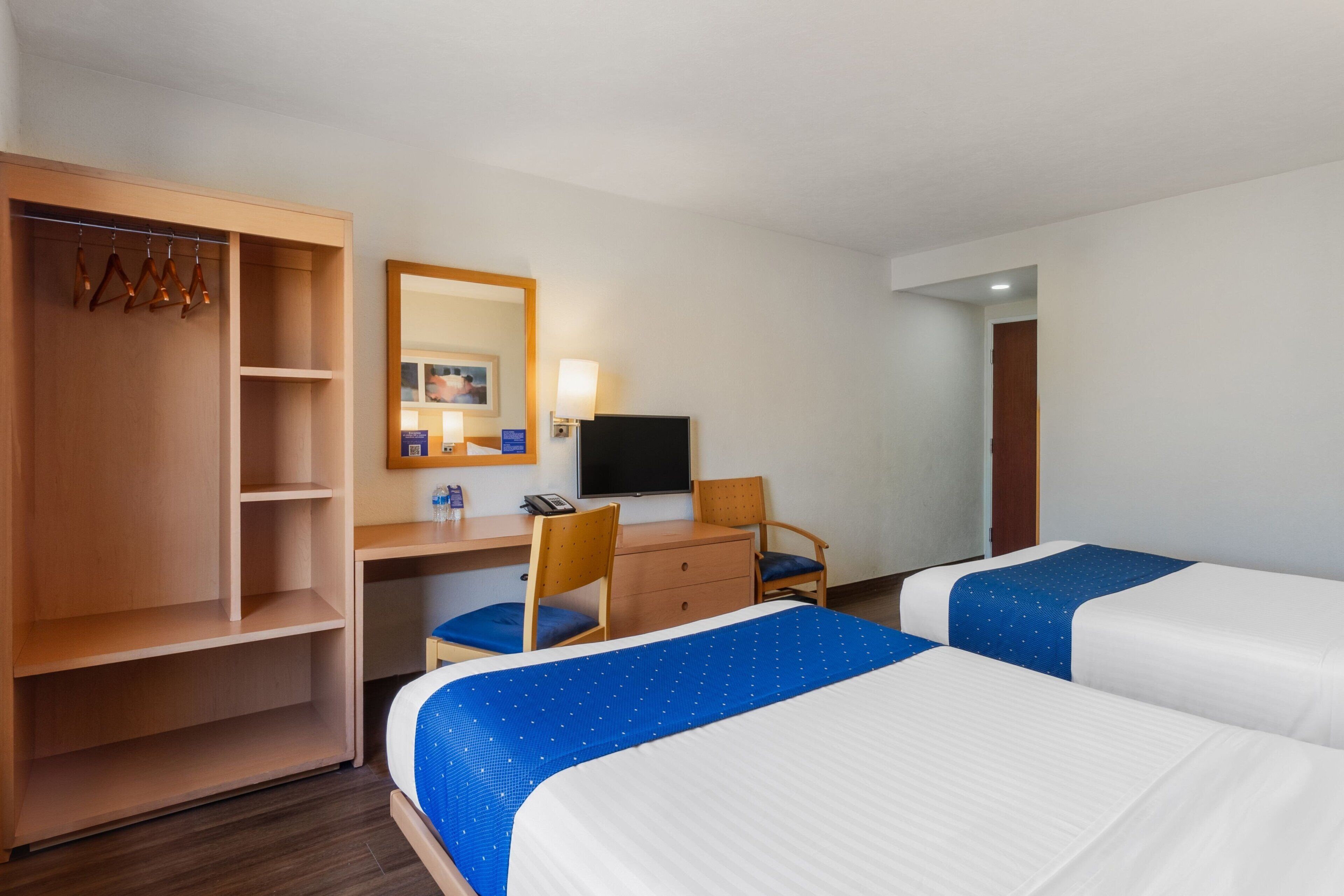 Habitación de hotel con dos camas, un escritorio con silla, un armario, un televisor y un espejo.