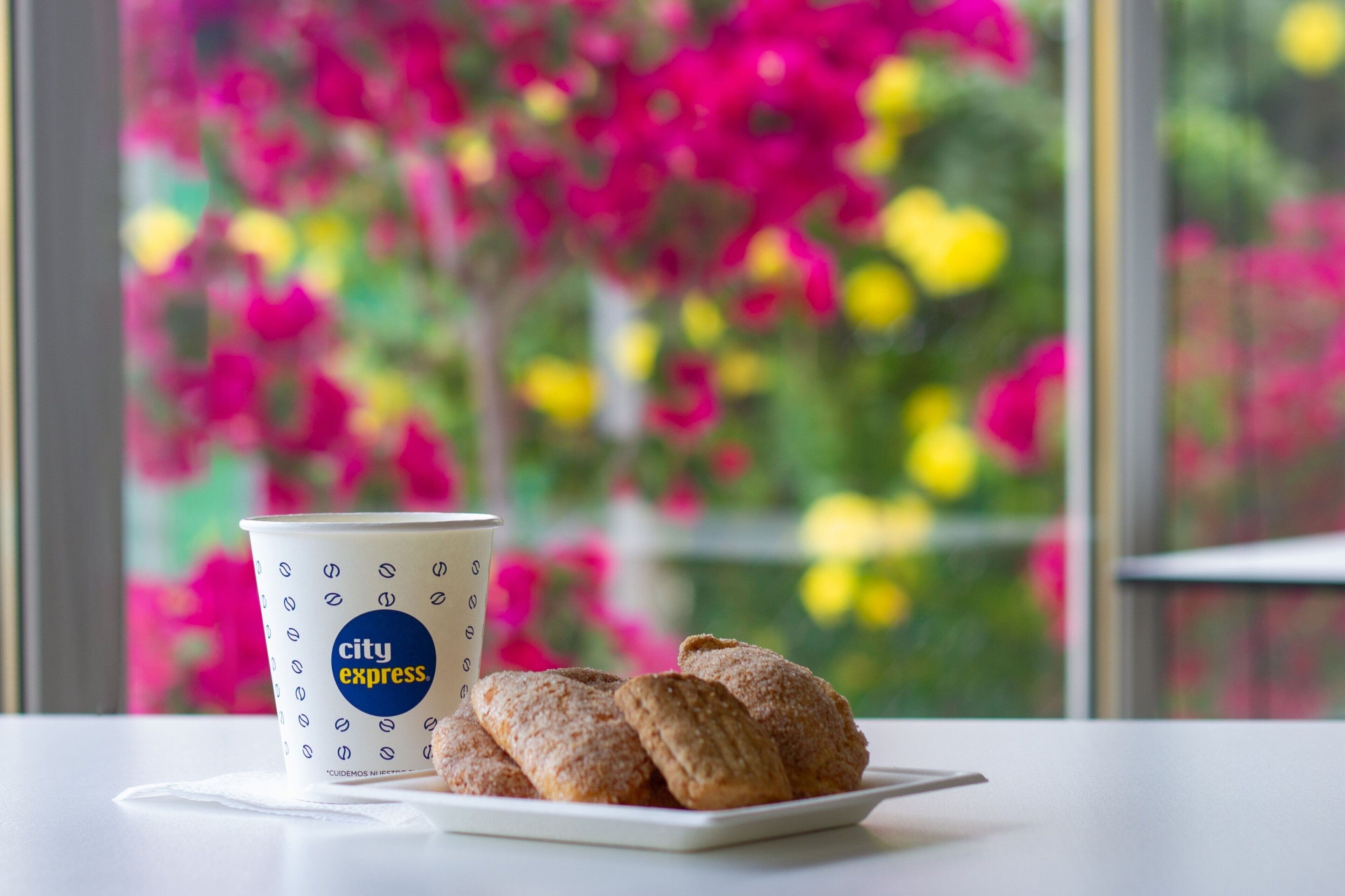 Una taza con el logotipo de City Express y un plato con pasteles sobre una mesa con vista a la ventana.