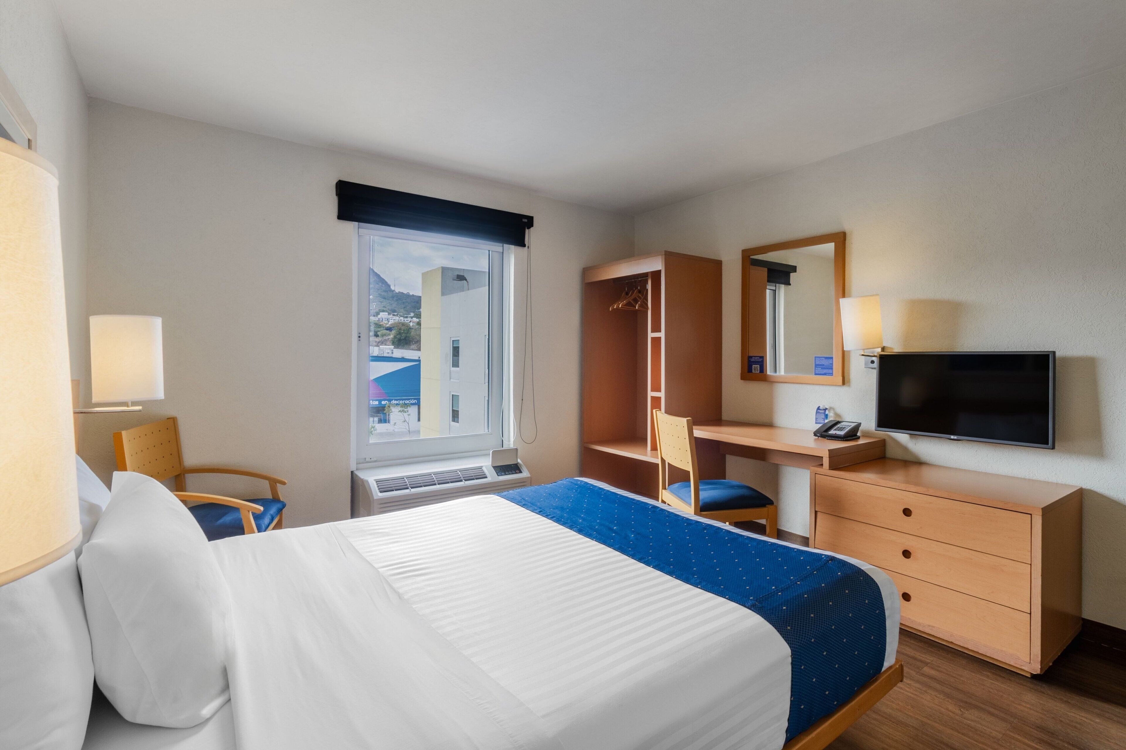 A hotel room with a bed, a desk, a chair, a TV, and a window with a view of buildings.