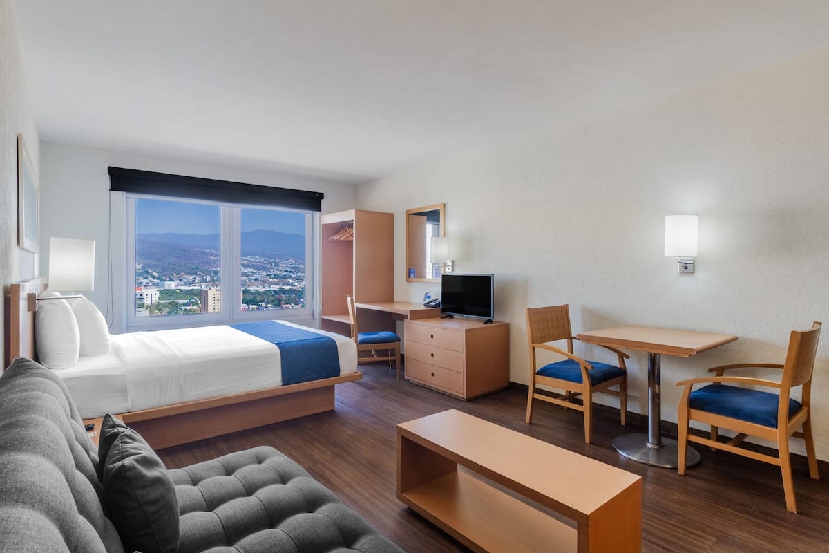 A hotel room with a bed, a desk, a chair, a sofa, and a view of a cityscape.