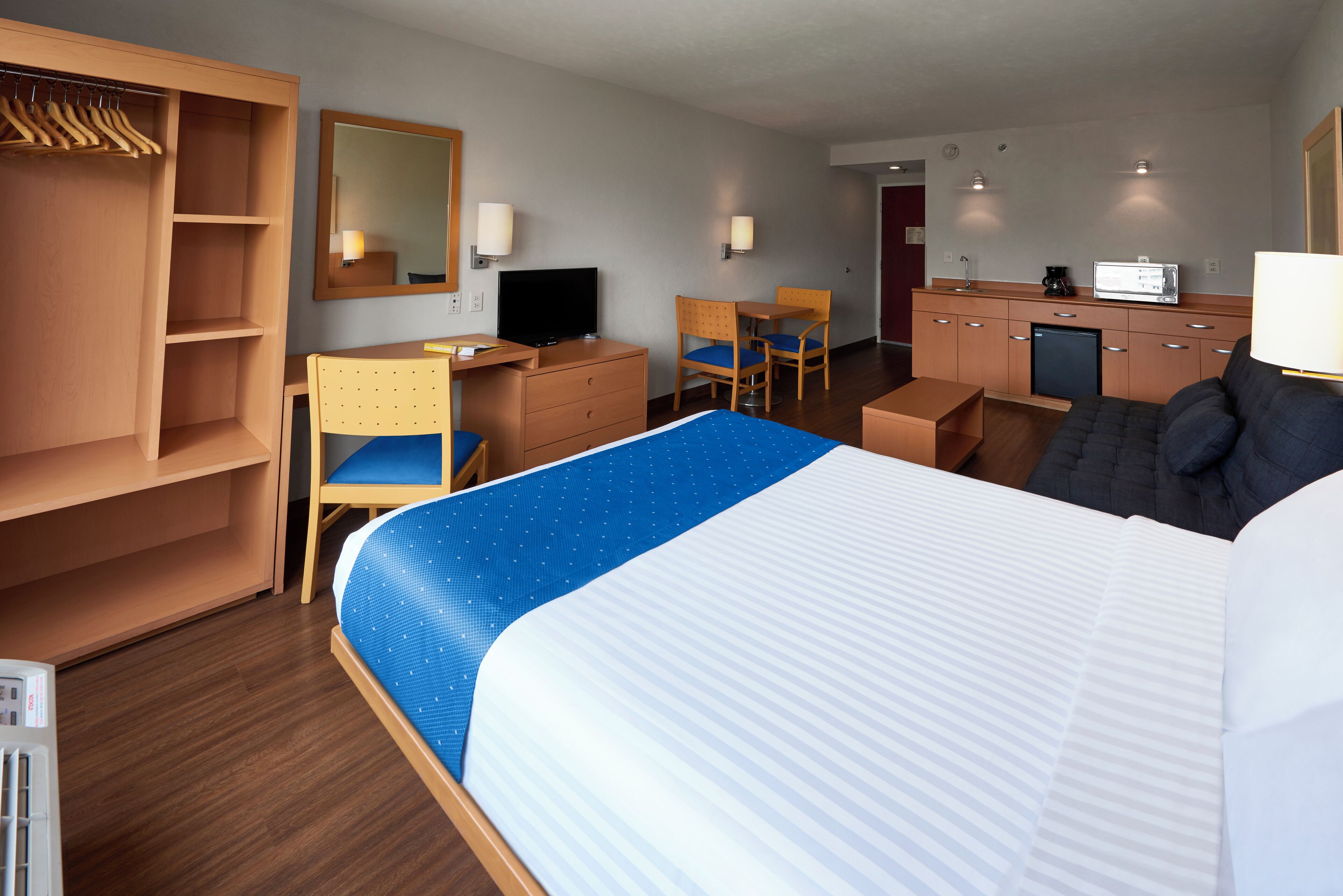 Quarto de hotel com cama, escrivaninha com cadeira, armário, televisão e cozinha compacta.