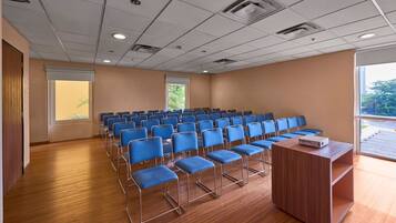 A conference room with rows of blue chairs, a wooden podium, and a large window with a view of greenery.