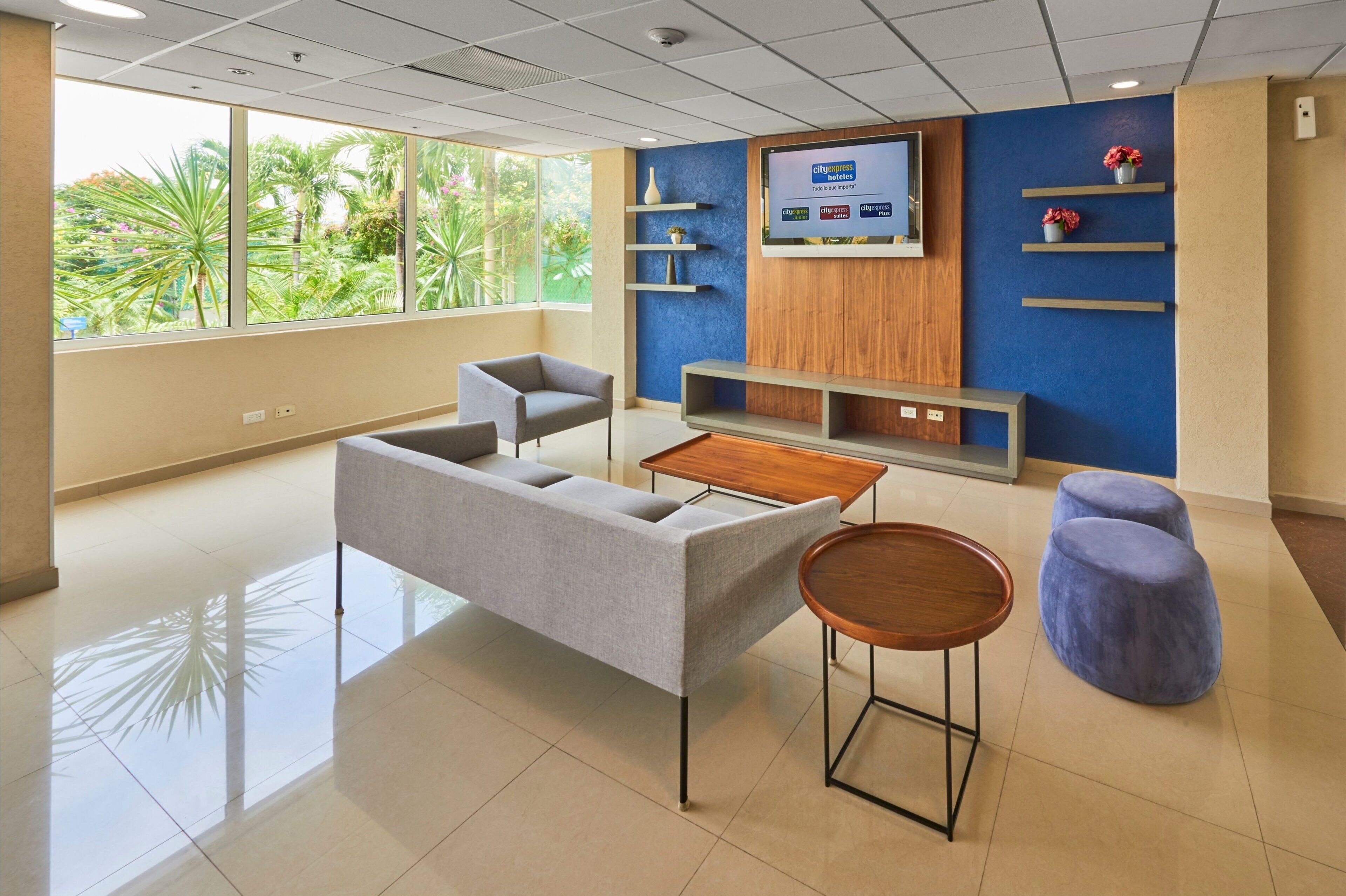 Un espace d’attente moderne comprenant un canapé, des fauteuils, une petite table et un écran de télévision diffusant des informations.