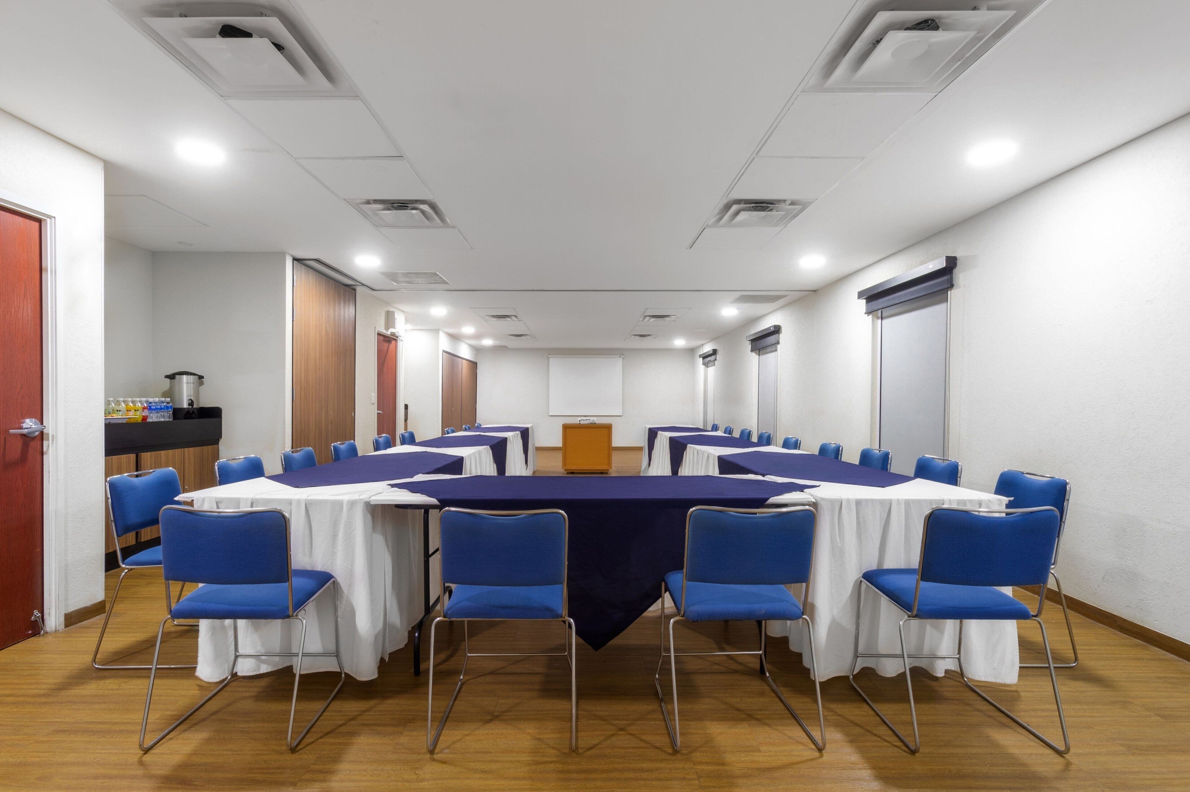 A conference room with a long table, blue chairs, white tablecloths, and a projector screen.