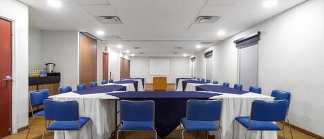 A conference room with a long table, blue chairs, white tablecloths, and a projector screen.
