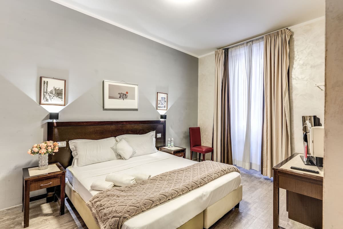 A hotel room with a large bed, a bedside table with a lamp, a chair, and a desk with a computer.