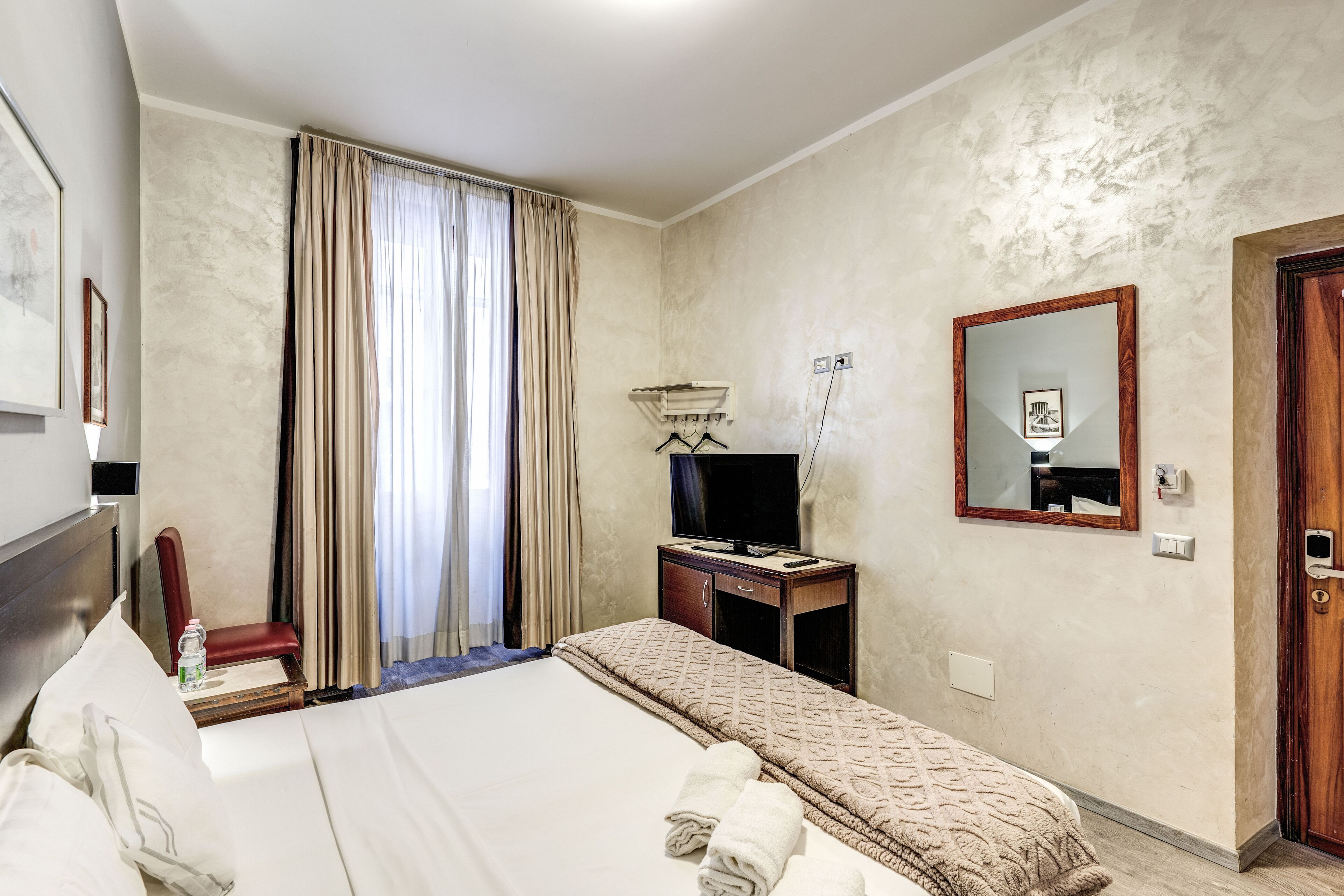 A hotel room with a bed, a television on a wooden stand, a mirror, and a door.
