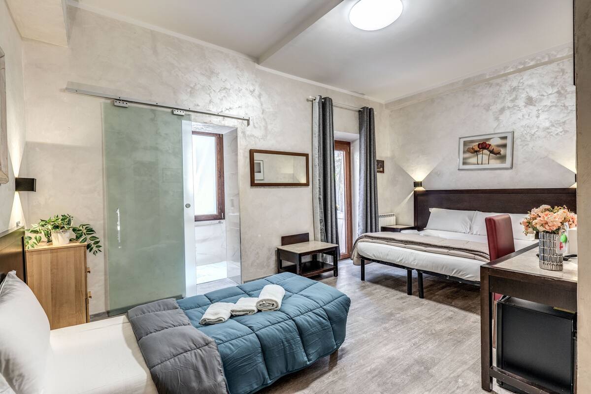 A hotel room with a bed, a desk, a chair, a small table, a cabinet, and a glass shower enclosure.