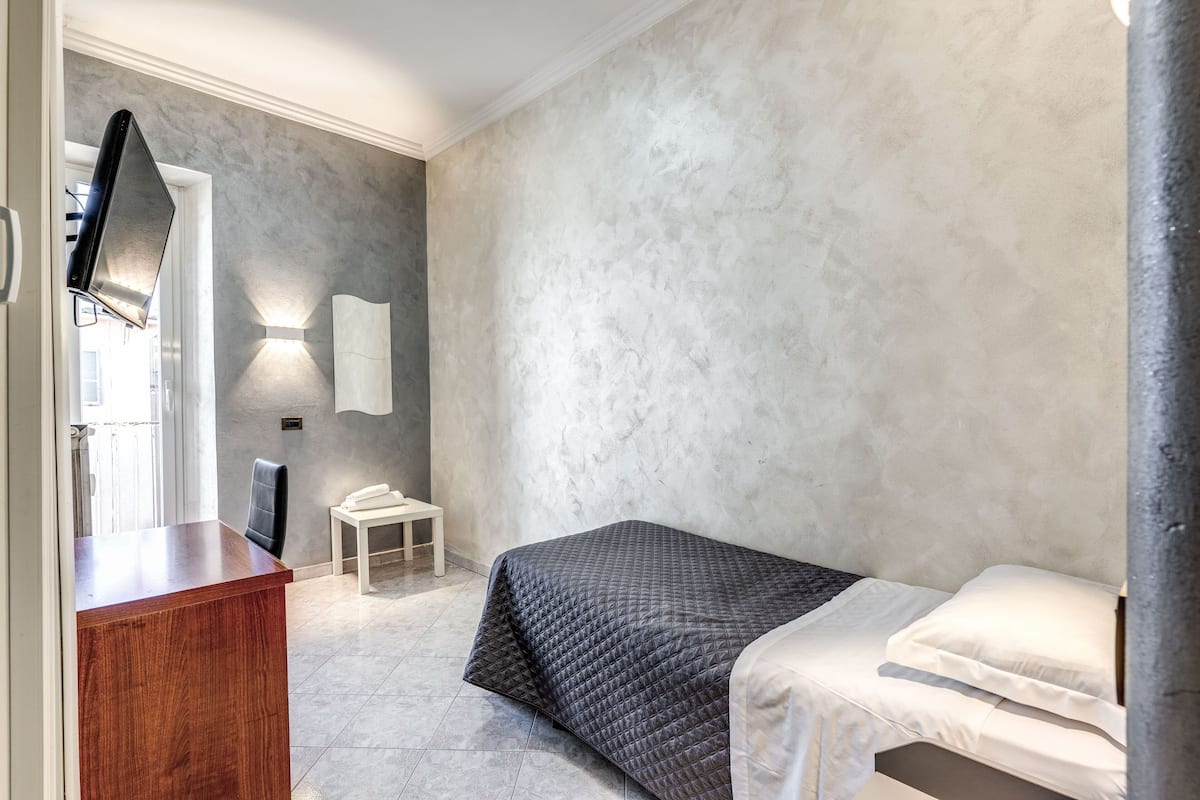 A bedroom with a bed, a desk, a chair, a TV mounted on the wall, and a wall-mounted light fixture.