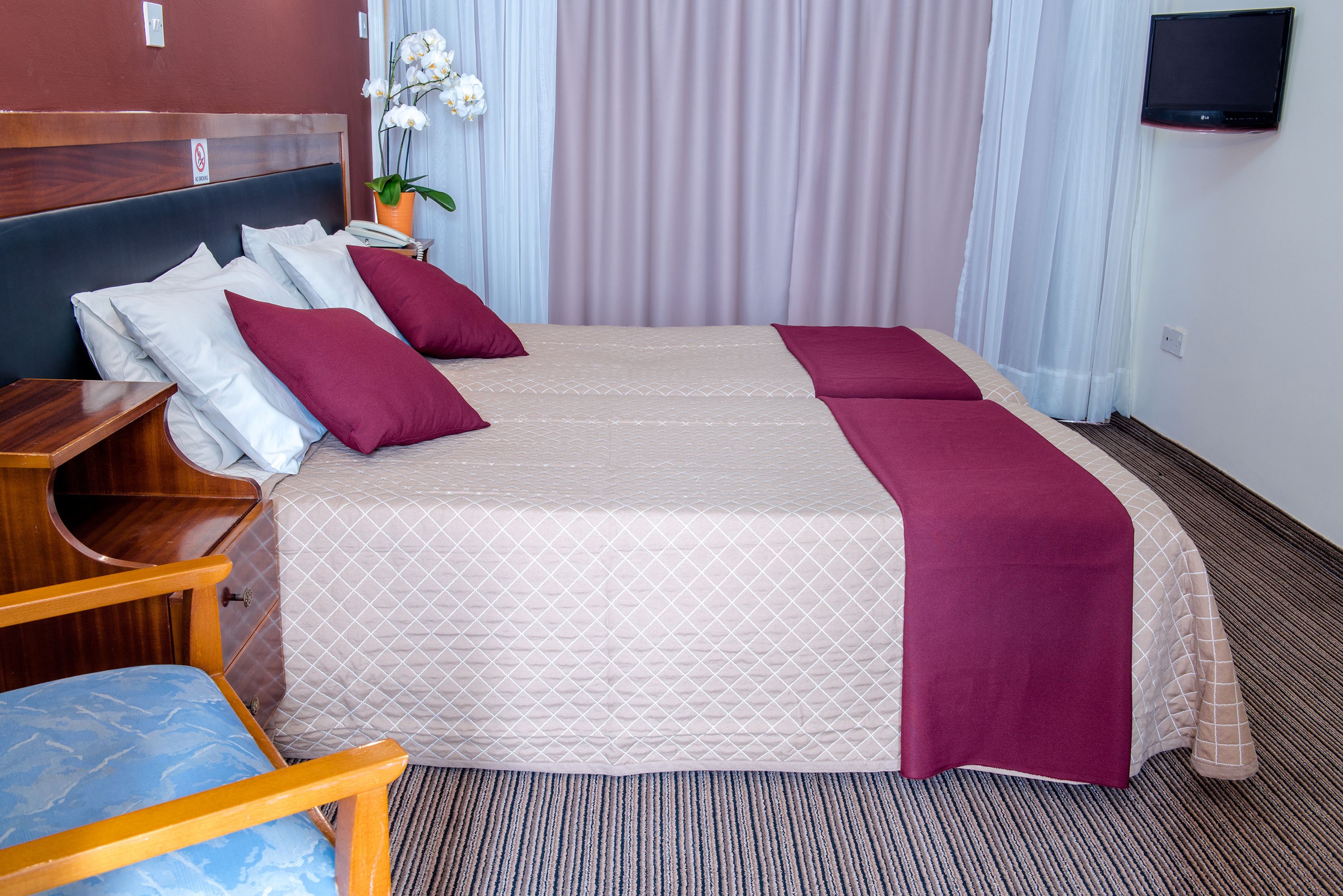 A hotel room with a large bed, two pillows, a wooden nightstand, a chair, a TV, and a potted plant.