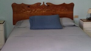 Un lit avec une tête de lit en bois, un oreiller bleu et une couvre-lit blanche à jupe. Il y a deux tables de chevet avec des lampes et un interrupteur fixé au mur.