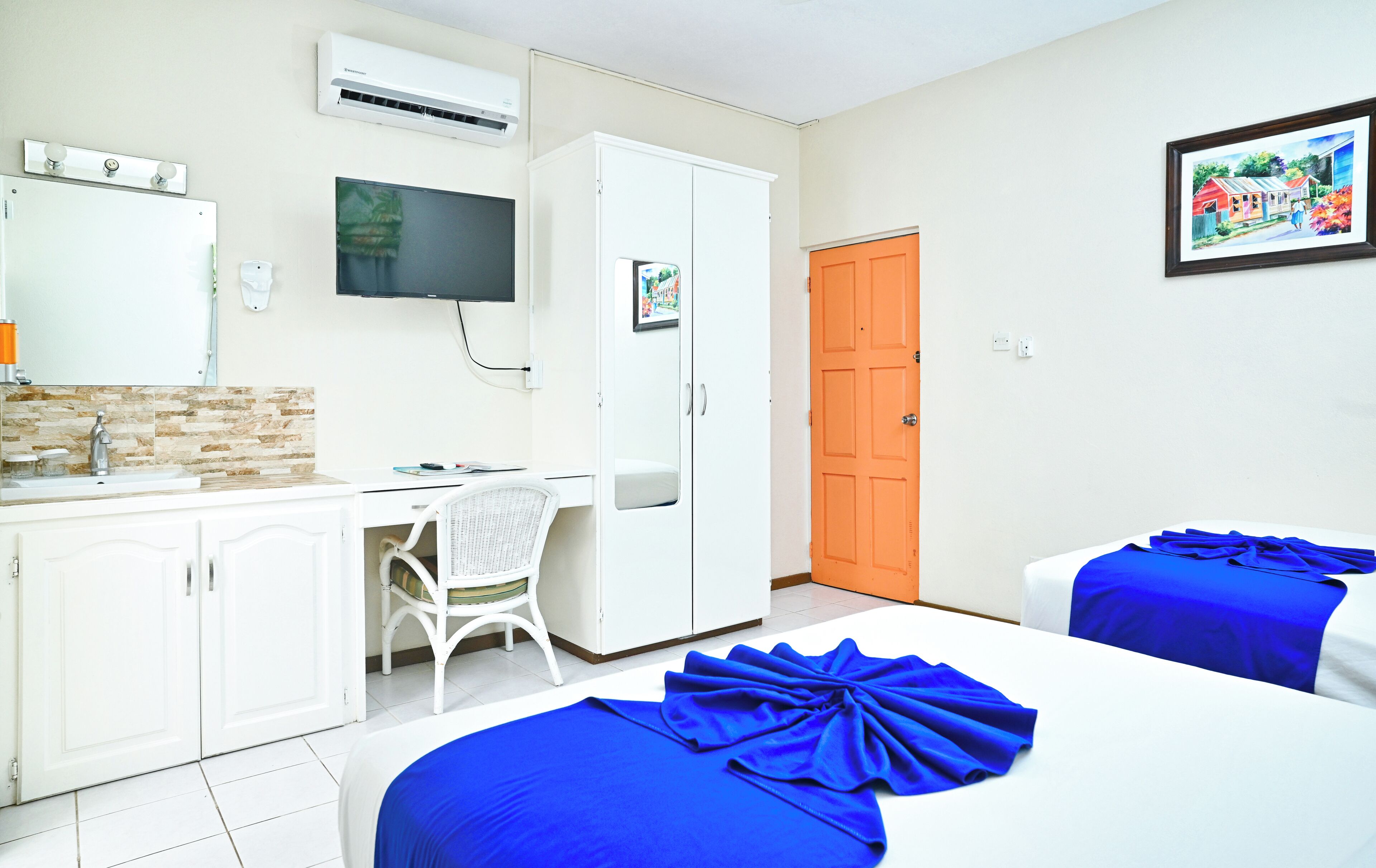 A room with a desk, a television, a wardrobe, an orange door, and two beds with blue bedspreads.