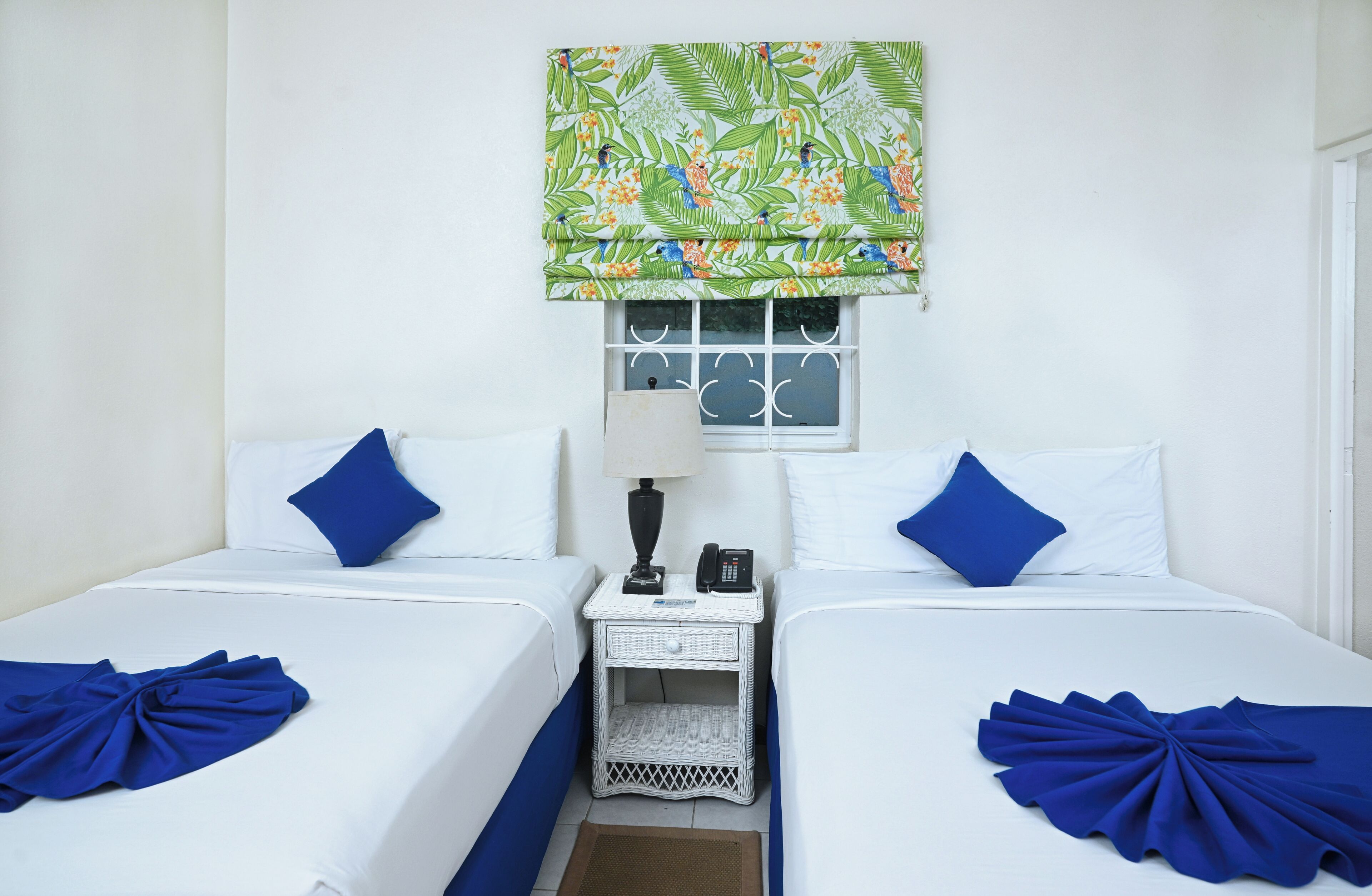 Habitación de hotel con dos camas, cada una con ropa blanca y almohadas azules. Hay una ventana con un alzaplastón estampado con hojas tropicales, una mesita de noche pequeña con una lámpara y un teléfono, y una toalla decorativa azul sobre cada cama.