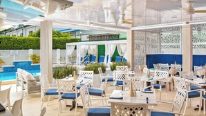An outdoor dining area with white lattice chairs and tables, a pool in the background, and a view of a building across the pool.
