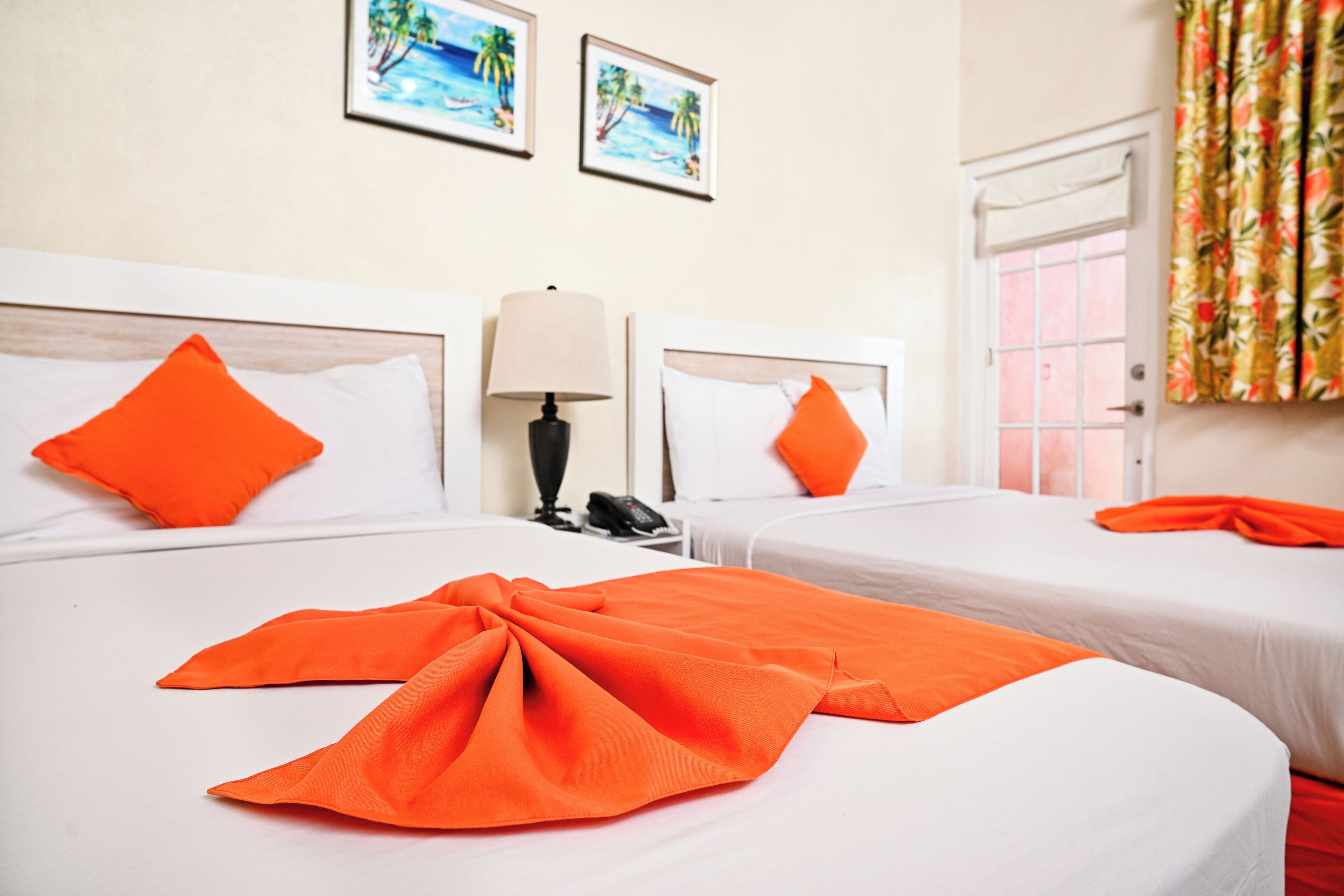Habitación de hotel con dos camas, cada una con ropa blanca y almohadas naranjas. Hay dos cuadros en la pared, una lámpara en la mesita de noche y una ventana con cortinas.