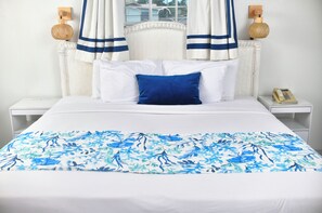 A neatly made bed with a floral bedspread, white bedding, and a blue pillow. There are two bedside tables with lamps and a telephone. - Melbourne Inn (St. Lawrence Gap)
