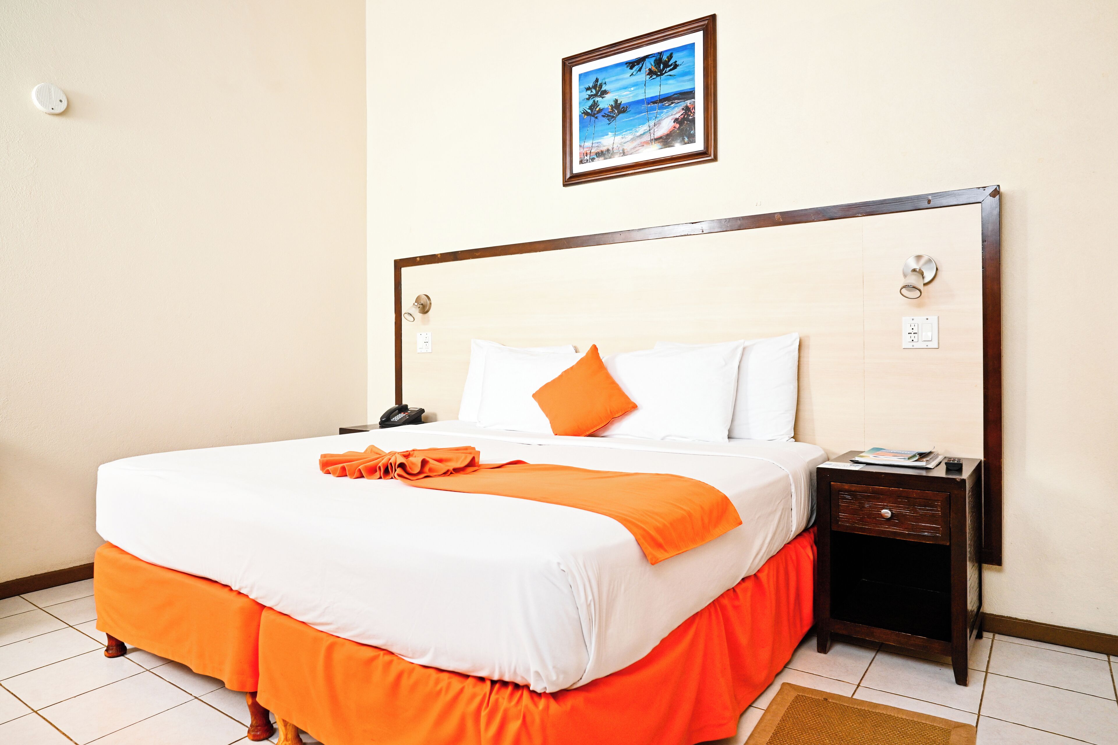 Una cama bien hecha con ropa de cama blanca y naranja, una mesita de noche con una lámpara y un cuadro en la pared.