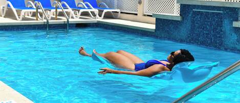 A pool with clear blue water, a person floating on a swim board, and blue lounge chairs along the poolside.