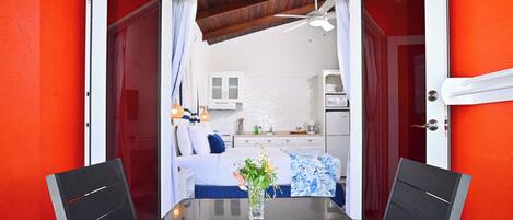 A room with a glass table and chairs, a white bed with blue and white bedding, and a kitchen area visible through an open door.