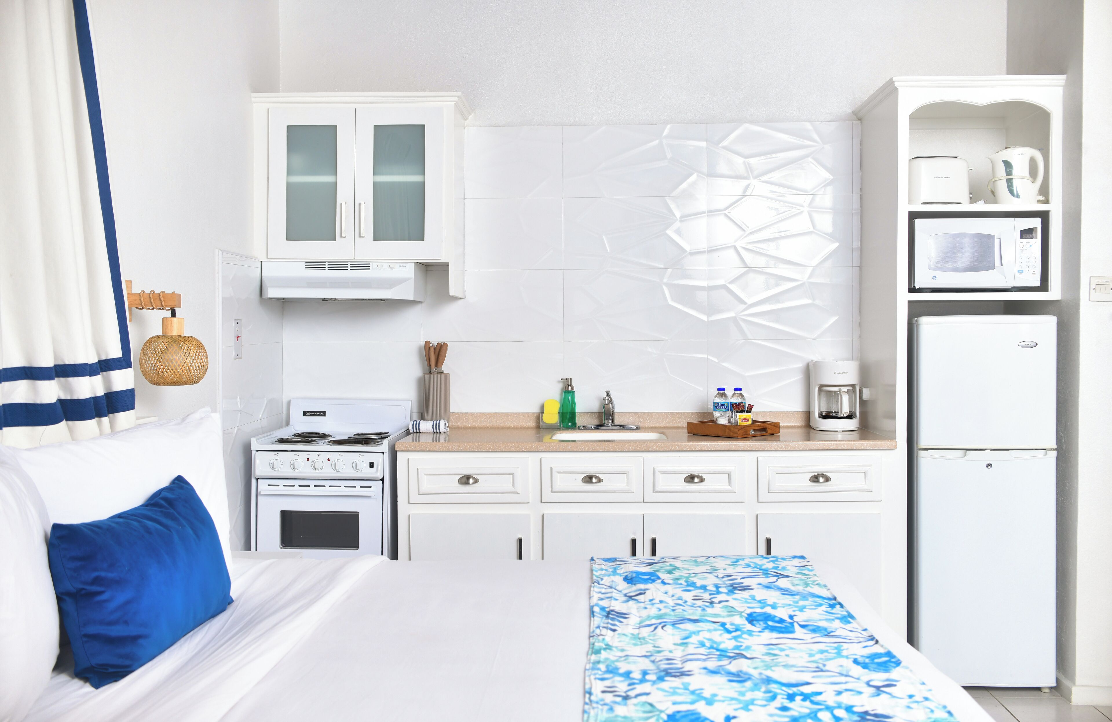 A hotel room with a bed, a kitchenette with a stove, microwave, and refrigerator, a sink, and a decorative wall piece.