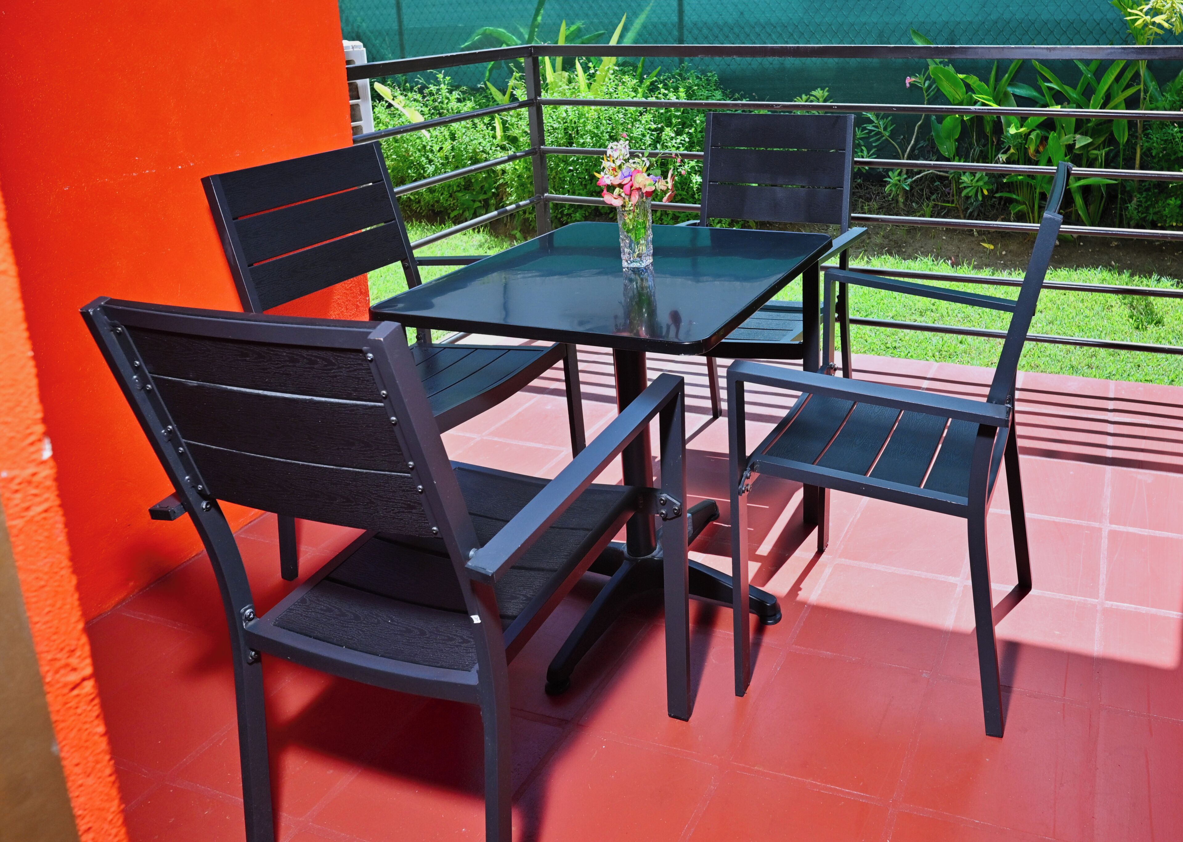 Un patio con una mesa y sillas, un florero con flores, y una cerca verde al fondo.