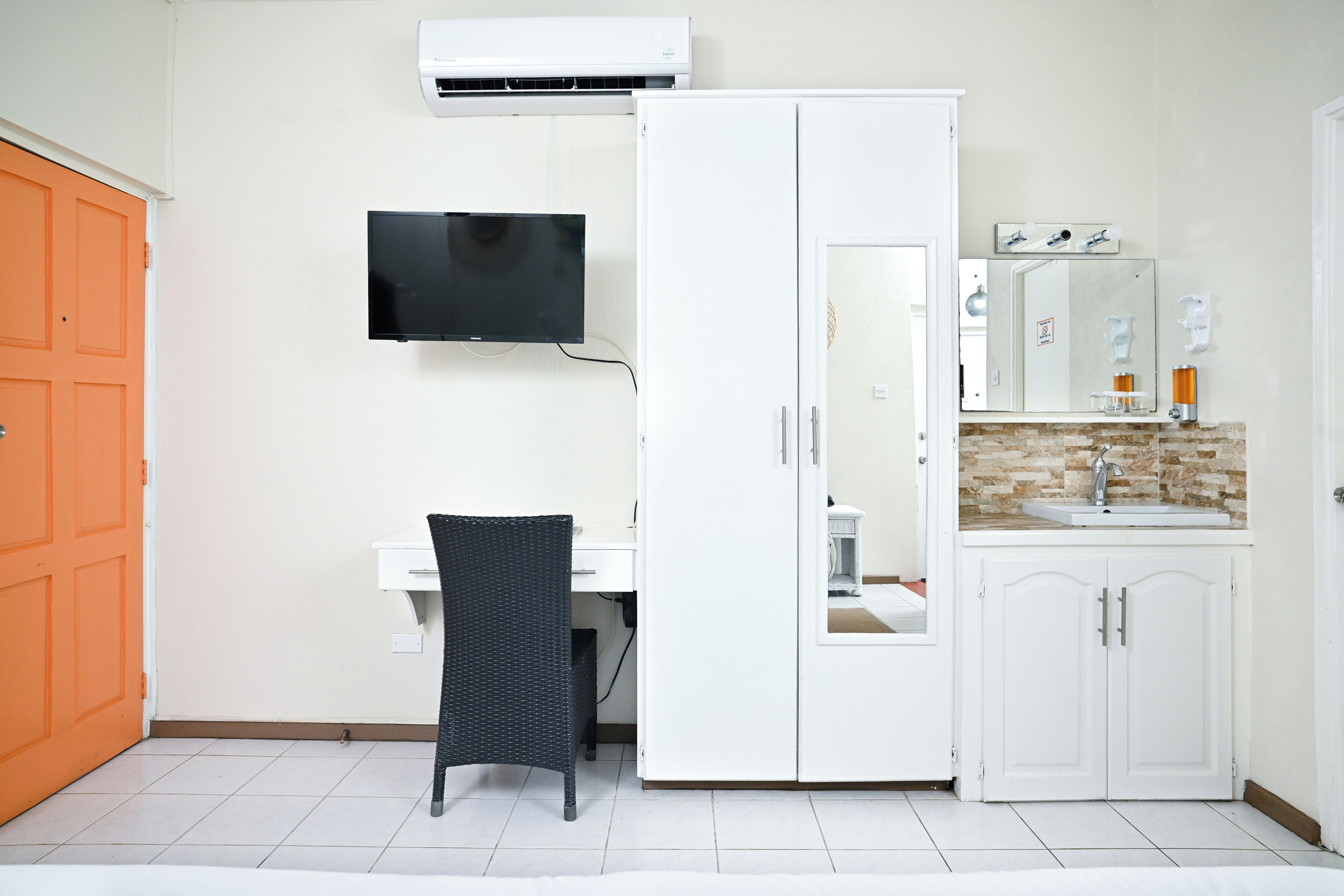 Een kamer met een witte kast, een aan de muur bevestigde televisie, een bureau met stoel, en een badkamer met wastafel en spiegel.