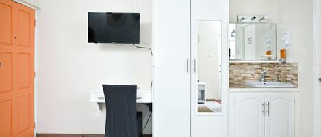 Een kamer met een witte kast, een aan de muur bevestigde televisie, een bureau met stoel, en een badkamer met wastafel en spiegel.
