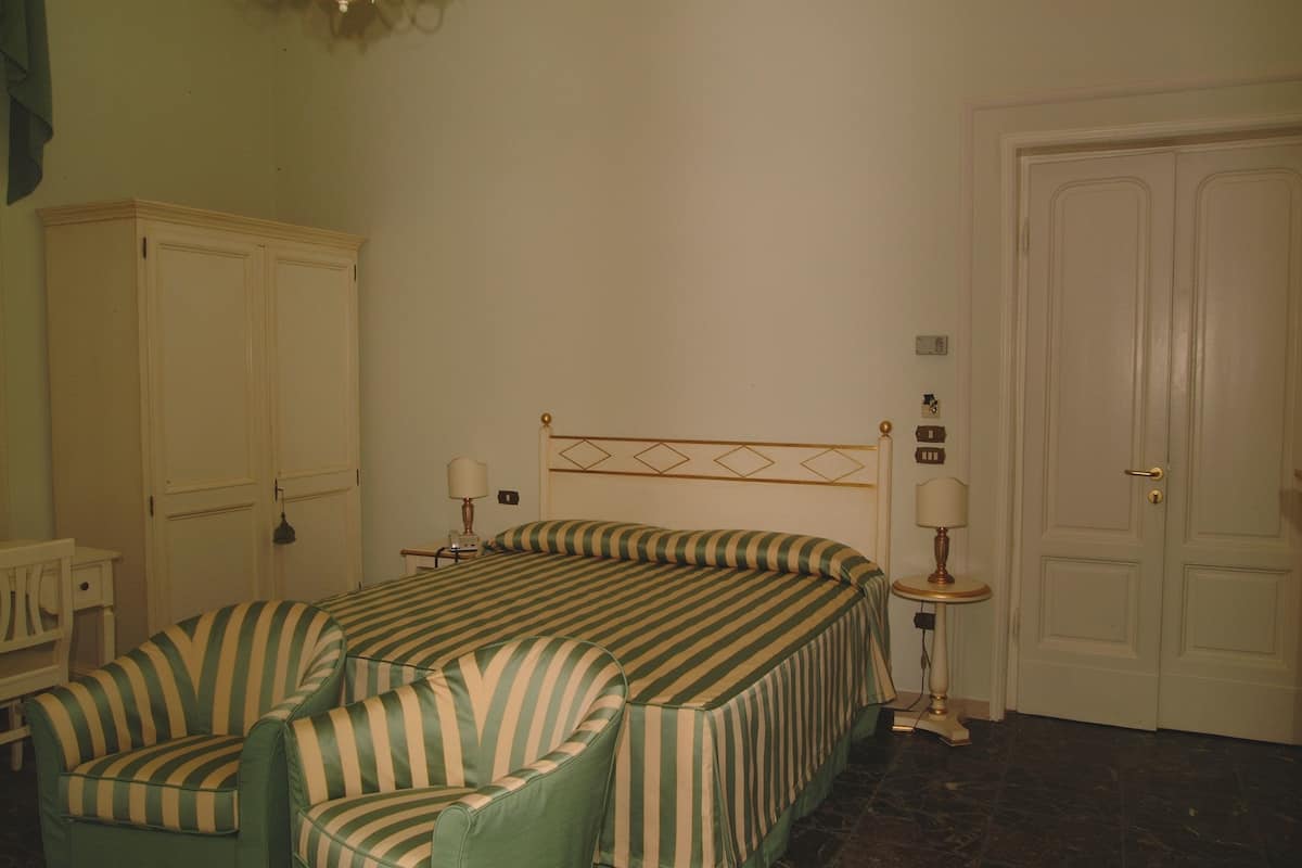 Ein Zimmer mit einem Bett, zwei gestreifte Sessel, einem Nachttisch mit Lampen, einem Kleiderschrank und einer Tür.