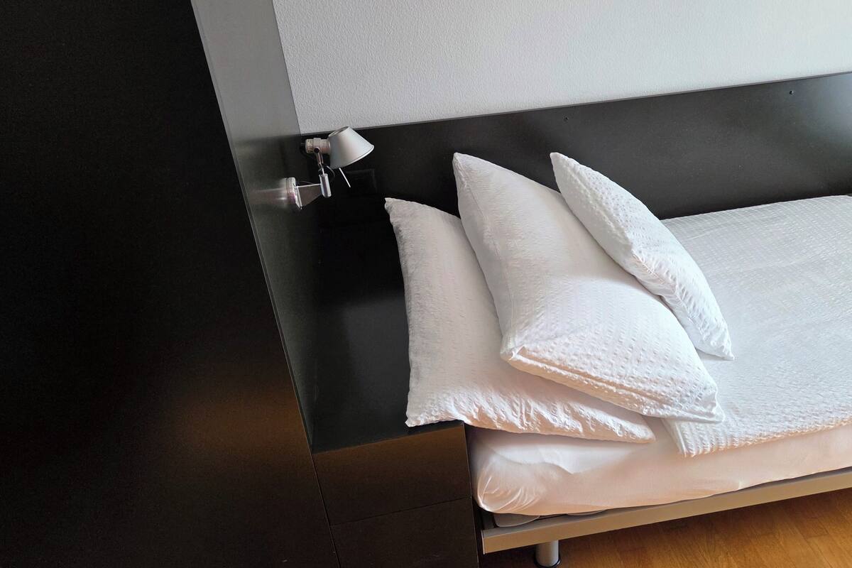 A neatly made bed with white bedding and pillows, a bedside lamp, and a dark headboard.