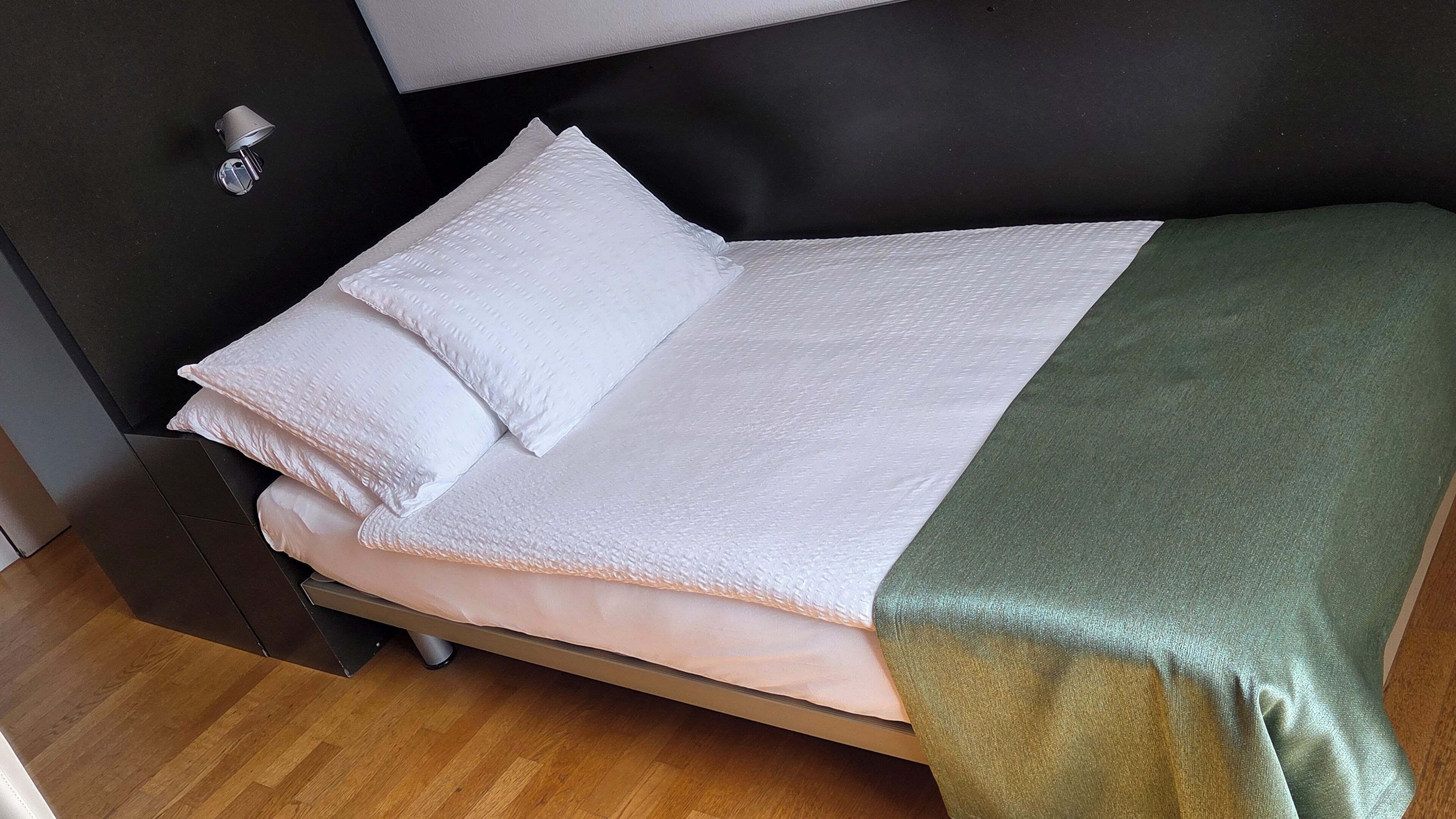 A single bed with a white mattress and pillows, a green blanket, and a black headboard.