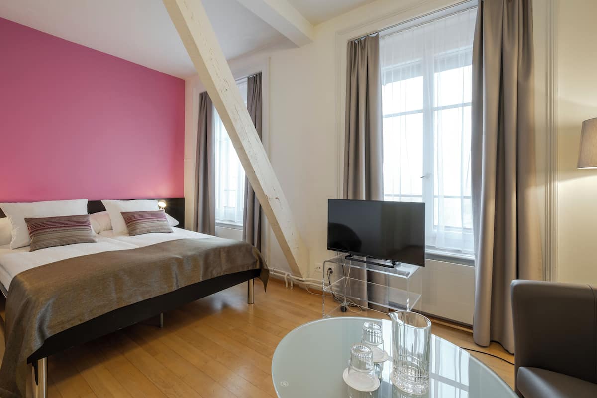 A modern hotel room with a bed, a TV, a glass table with glasses, and large windows with curtains.