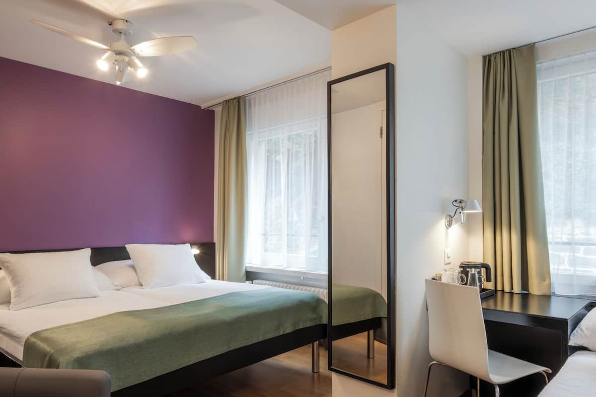 A hotel room with a bed, a desk, a chair, a mirror, and a ceiling fan.