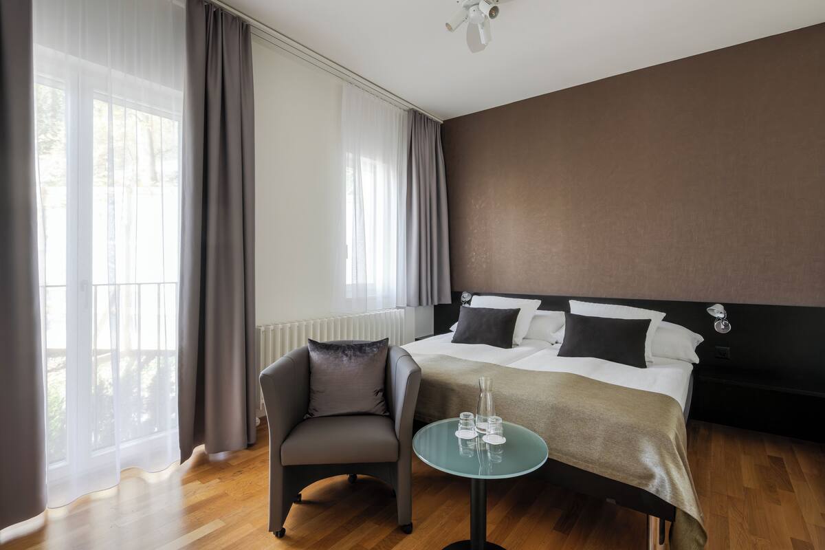 A hotel room with a bed, a chair, a small table, and a view of the outdoors.