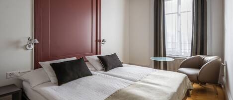 A hotel room with two beds, a red door, a window with curtains, a small round table, and a leather chair.