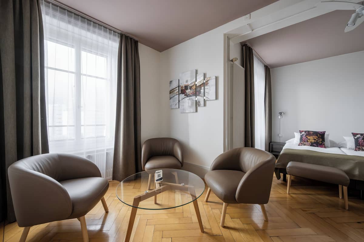 A modern room with a glass coffee table, two leather armchairs, a bed with floral pillows, and a large window with sheer curtains.