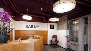 A hotel reception area with a wooden reception desk, a black sign reading "KARL HOTEL LUZERN," a modern chair with a pink cushion, and a small table with a potted plant.