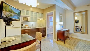 A modern kitchen with a flat-screen TV, white cabinets, a microwave, and a dining area with a chair and table.