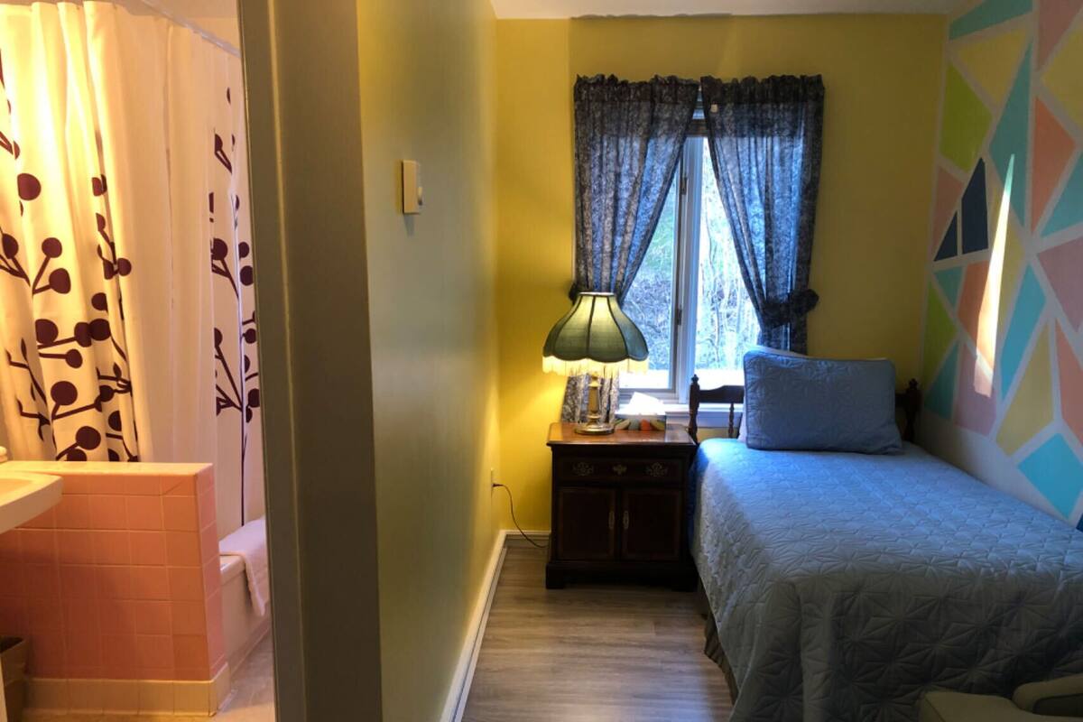 A bedroom with a bed, a nightstand, a lamp, a window with curtains, and a colorful wall design.