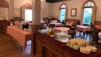 A dining area with multiple tables, each covered with a pink tablecloth. There are trays with plates, glasses, and a pitcher of water. The room has wooden floors, chandeliers, and framed pictures on the walls.