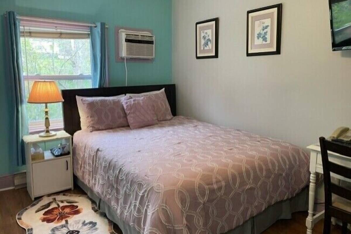 A bedroom with a bed, a nightstand, a lamp, an air conditioner, and framed pictures on the wall.