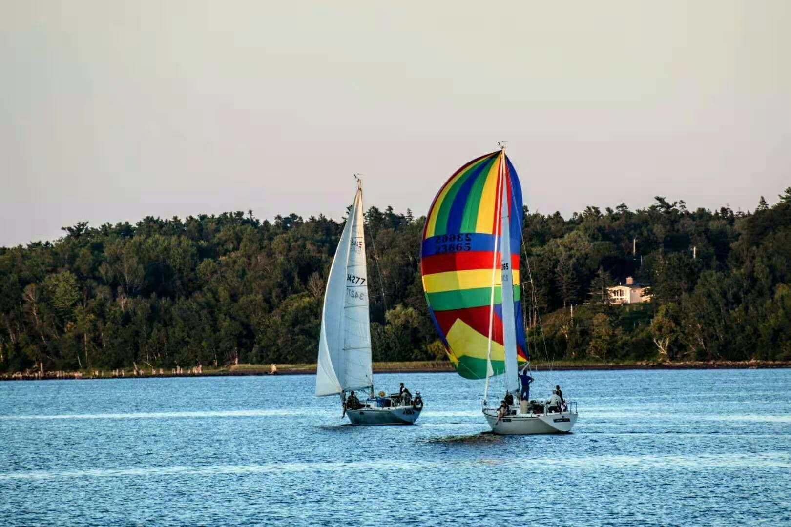 Two sailboats on a calm body of water with a forested shoreline in the background.