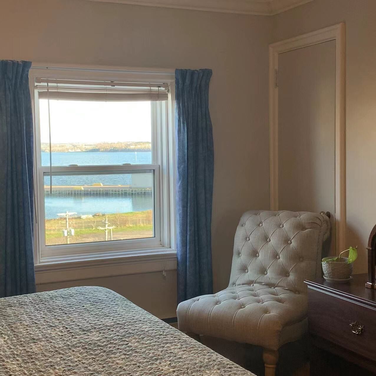Un dormitorio con cama, una silla y vista a un cuerpo de agua a través de una ventana con cortinas azules.