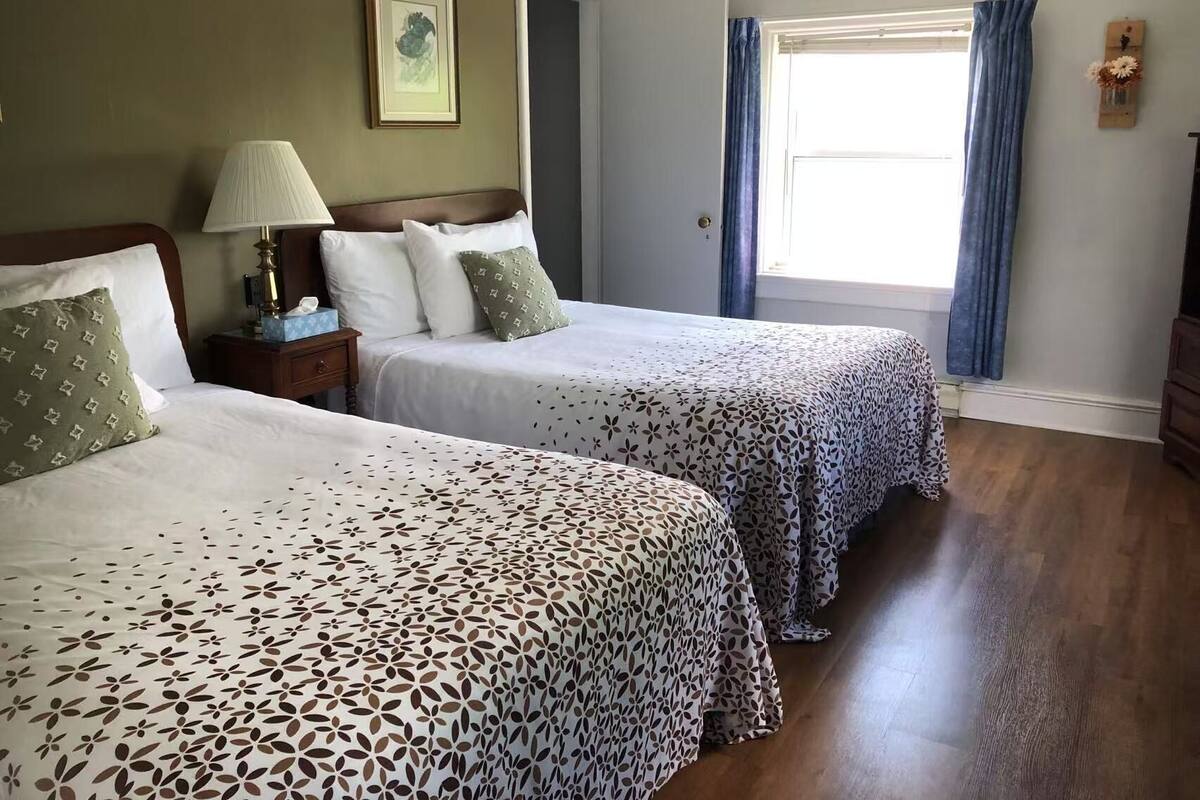 A hotel room with two beds, a nightstand, a lamp, a framed picture on the wall, and a window with curtains.