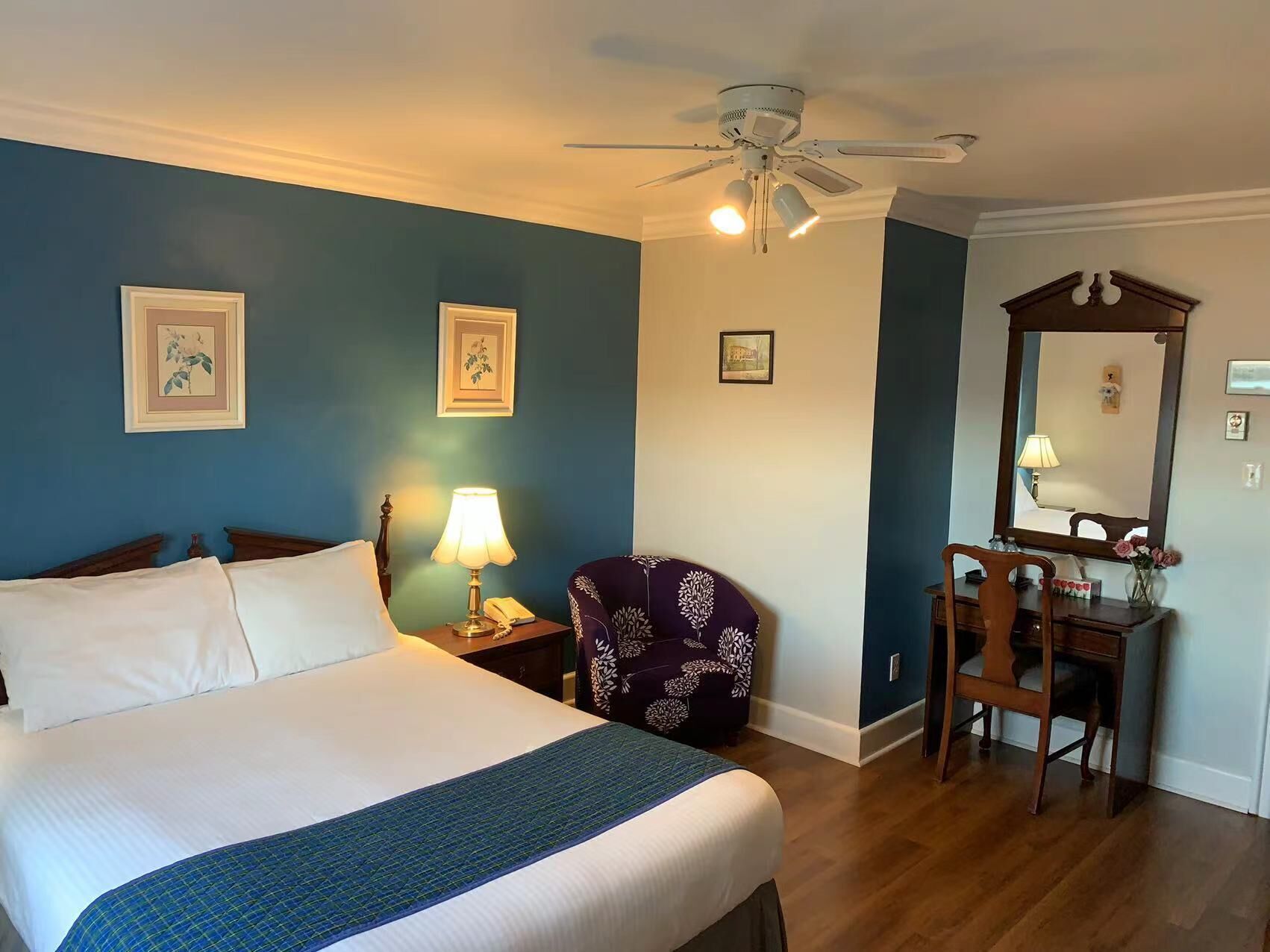 A hotel room with a bed, a desk with a chair, a ceiling fan, and framed pictures on the walls.