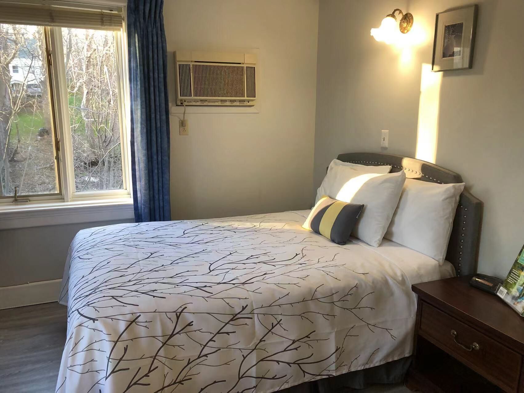 Un dormitorio con una cama, una mesita de noche, un aire acondicionado de pared y una ventana con cortinas.