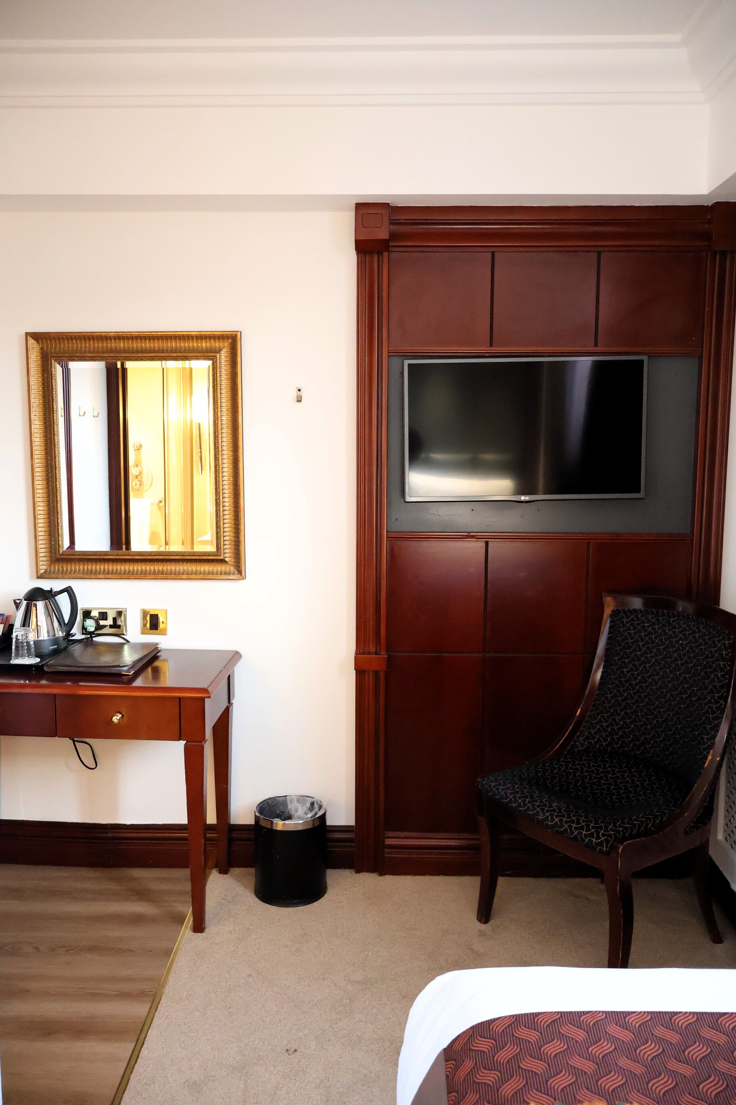 A hotel room with a bed, a desk with a coffee maker, a chair, a trash can, a mirror, and a TV mounted on the wall.