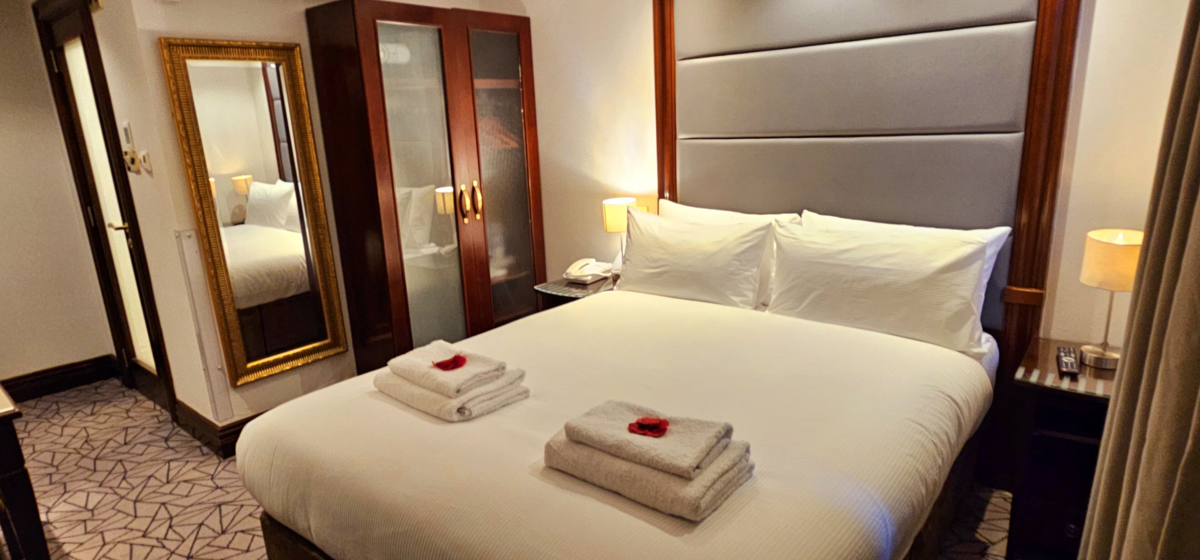 A hotel room with a large bed, a mirror, a wardrobe, and a bedside table with a lamp.